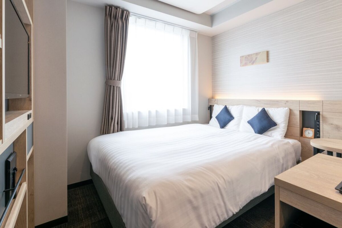 Une chambre d’hôtel avec un lit, une table de chevet avec un plateau, une horloge murale et une fenêtre avec des rideaux.