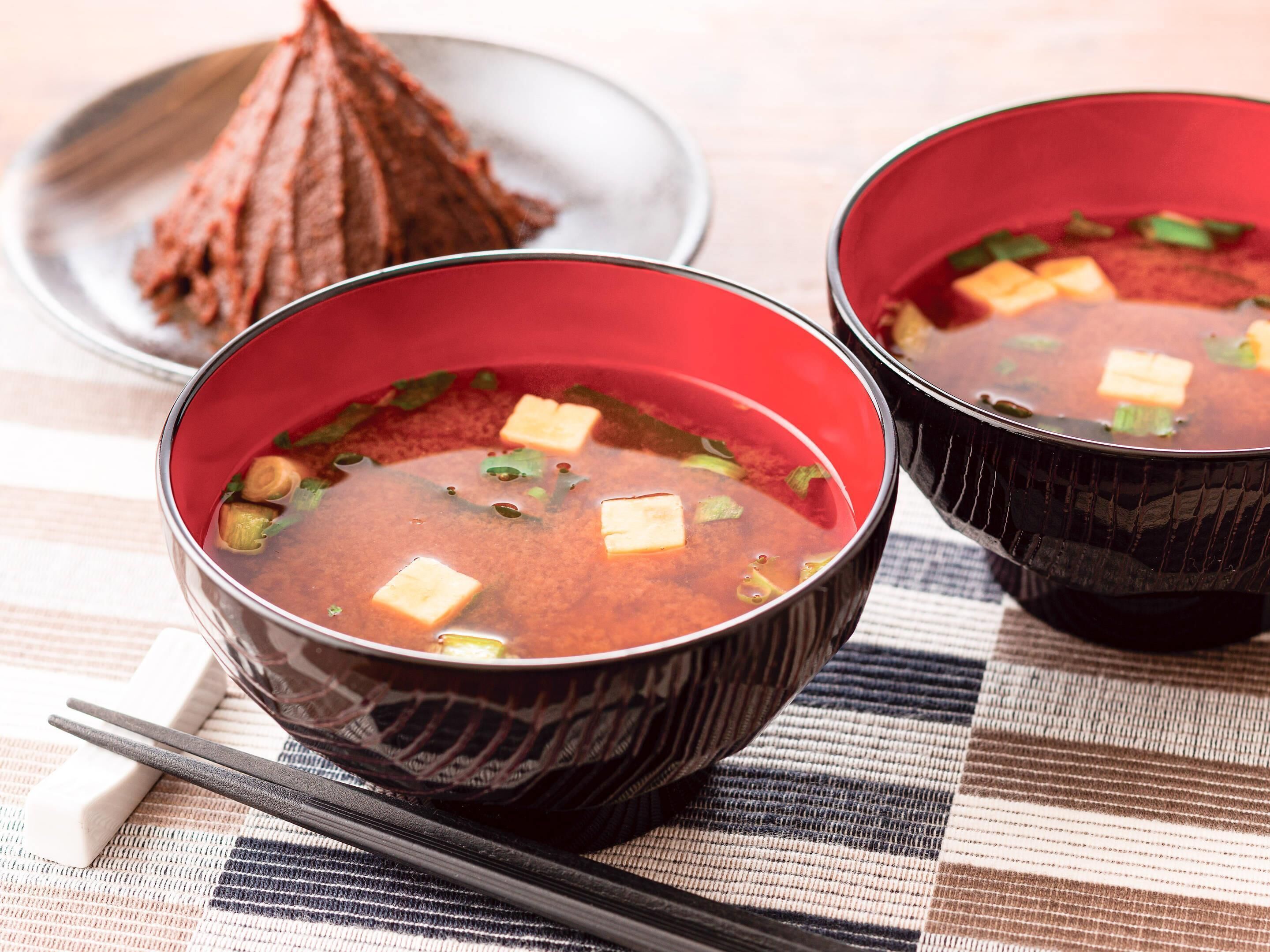 Two bowls of red soup with tofu cubes and green onions, accompanied by chopsticks and a white object on a checkered tablecloth.