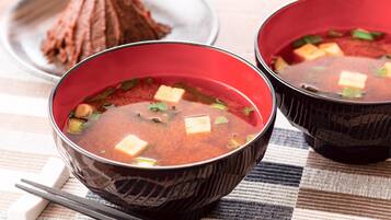 Two bowls of red soup with tofu cubes and green onions, accompanied by chopsticks and a white object on a checkered tablecloth.