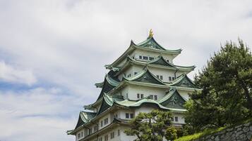 Un castillo japonés tradicional con varios niveles y tejados verdes, rodeado de árboles y un muro de piedra.