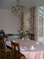 Comedor con mantel rosa, sillas de madera, lámpara de araña y cortinas con motivos florales.