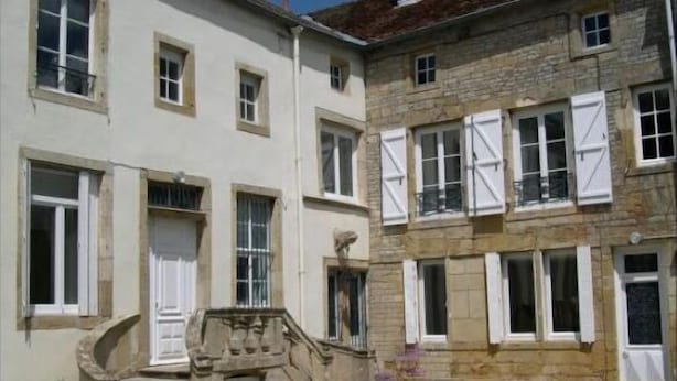 Un edificio de dos pisos con fachada de piedra y contraventanas blancas. Una escalera de piedra conduce a la entrada.