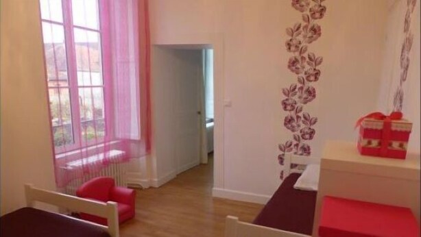 Quarto com duas camas, janela com cortinas rosa, decoração floral na parede e uma pequena mesa com uma caixa vermelha.