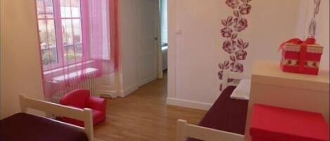 Una habitación con dos camas, una ventana con cortinas rosadas, un adorno floral en la pared y una mesita con una caja roja.