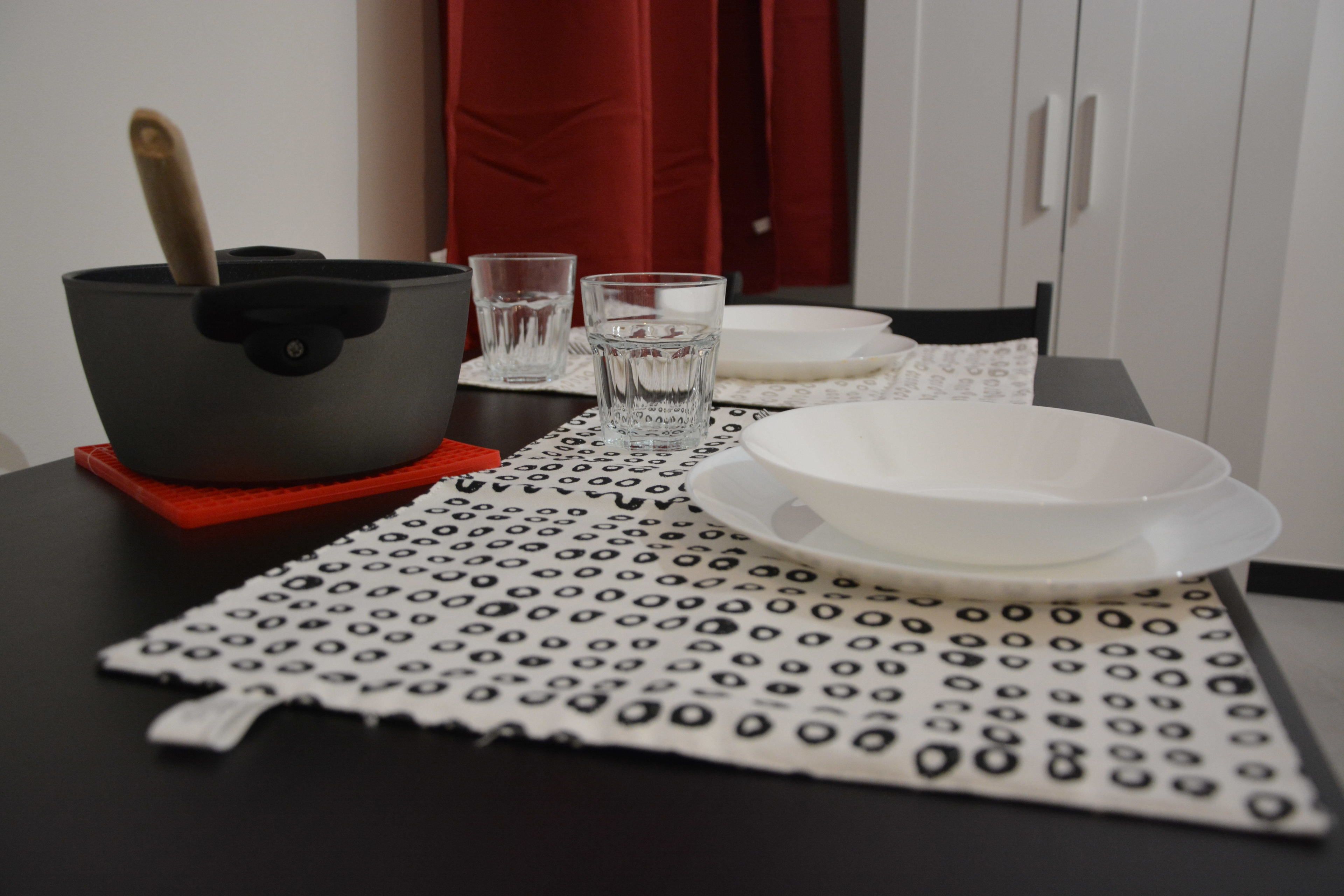 Une table dressée avec une nappe à motifs, une assiette blanche, un verre et un pot muni d’une anse.