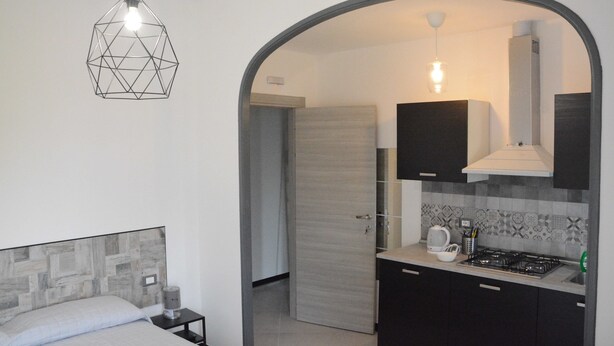 A modern bedroom with a geometric-patterned headboard, a bedside table with a lamp, and a large arched doorway leading to a kitchen with dark cabinets and a tiled backsplash. - Casa dei Nonni (Genoa)