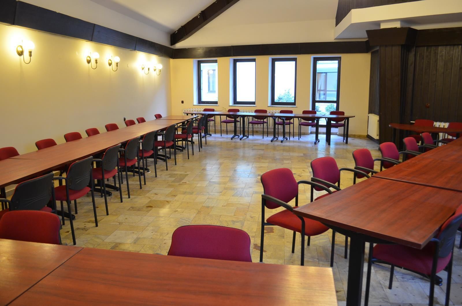 Un salón de conferencias con largas mesas y sillas rojas, ventanas amplias e iluminación empotrada en la pared.