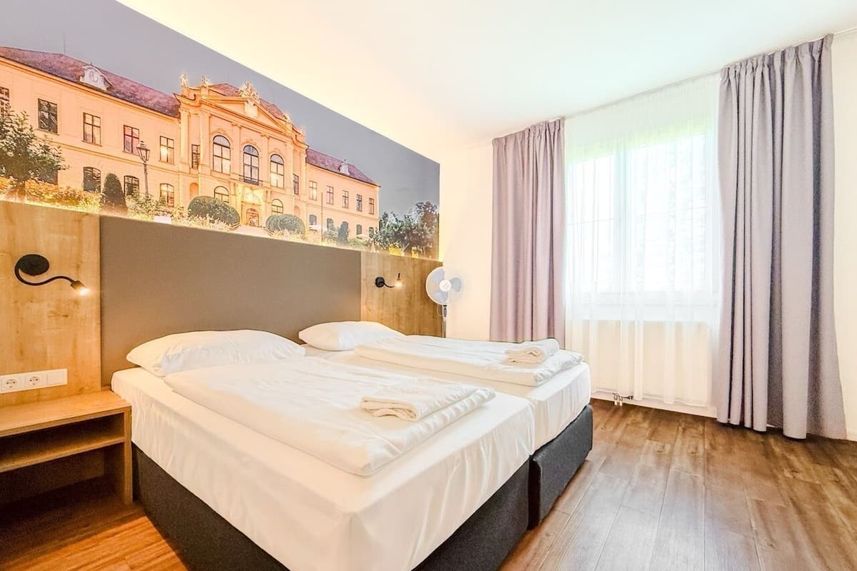 Quarto de hotel com duas camas, um mural de um edifício, um banco de madeira, uma mesinha de cabeceira e uma janela com cortinas.