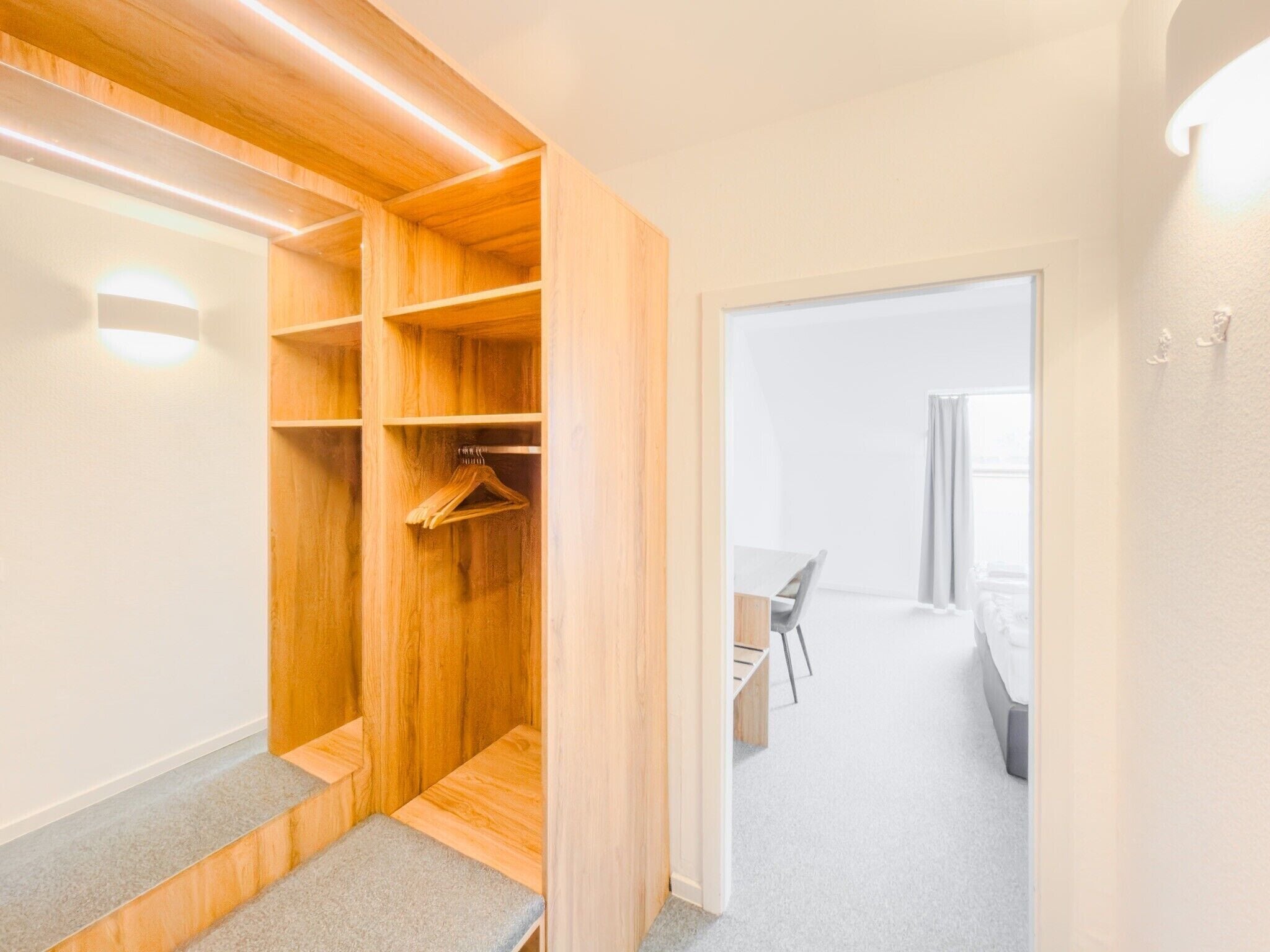 A walk-in closet with wooden shelving and a bench, leading to a bedroom with a bed and a window.