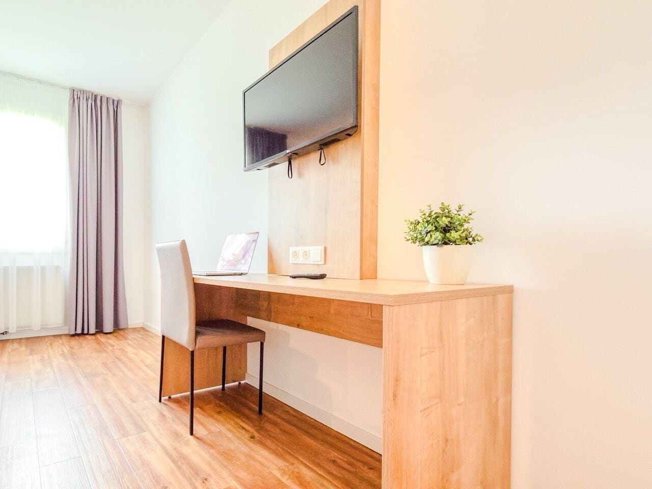 Una habitació moderna amb escriptori de fusta, una cadira, un ordinador portàtil, televisió penjada a la paret i una planta en test.