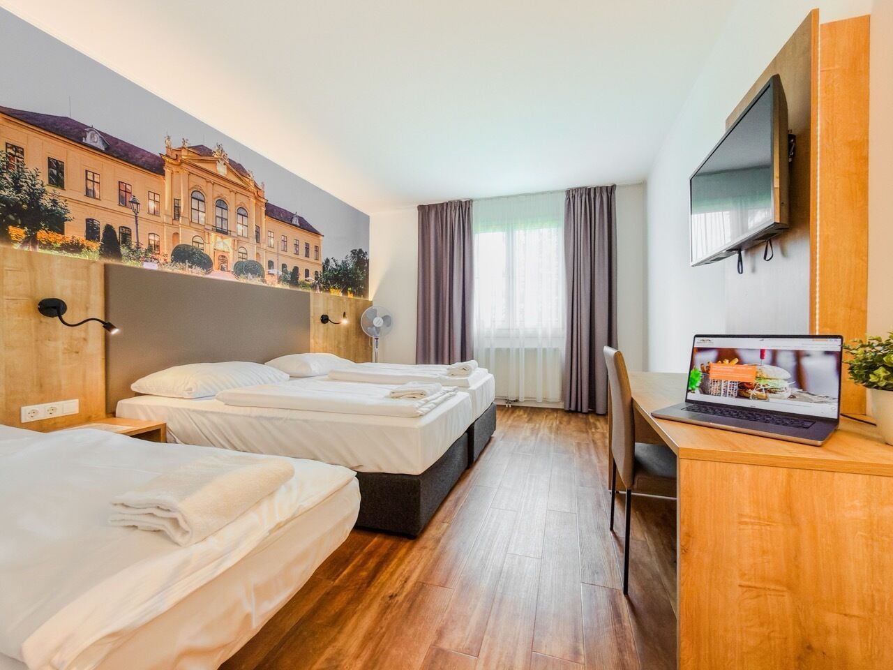 A hotel room with two beds, a desk with a laptop, a TV, and a mural of a building.