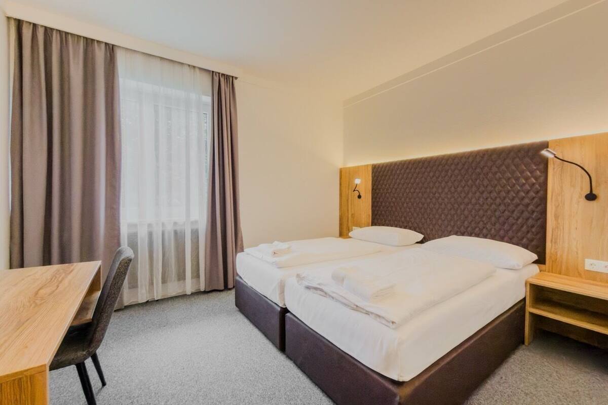 Quarto de hotel com cama, escrivaninha, cadeira e janela com cortinas.