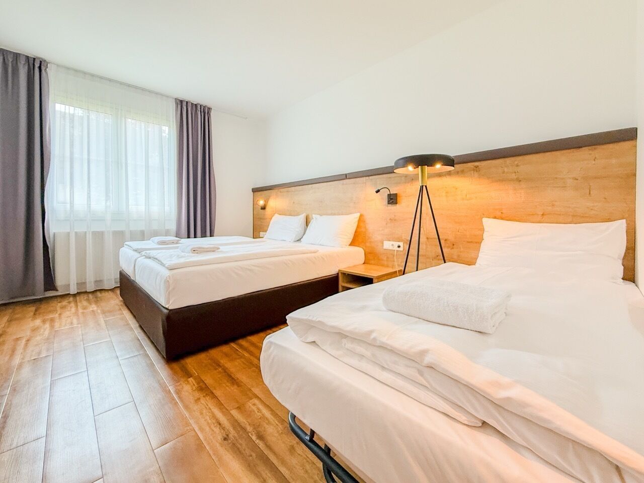 A hotel room with two beds, a wooden headboard, a bedside lamp, and a window with curtains.