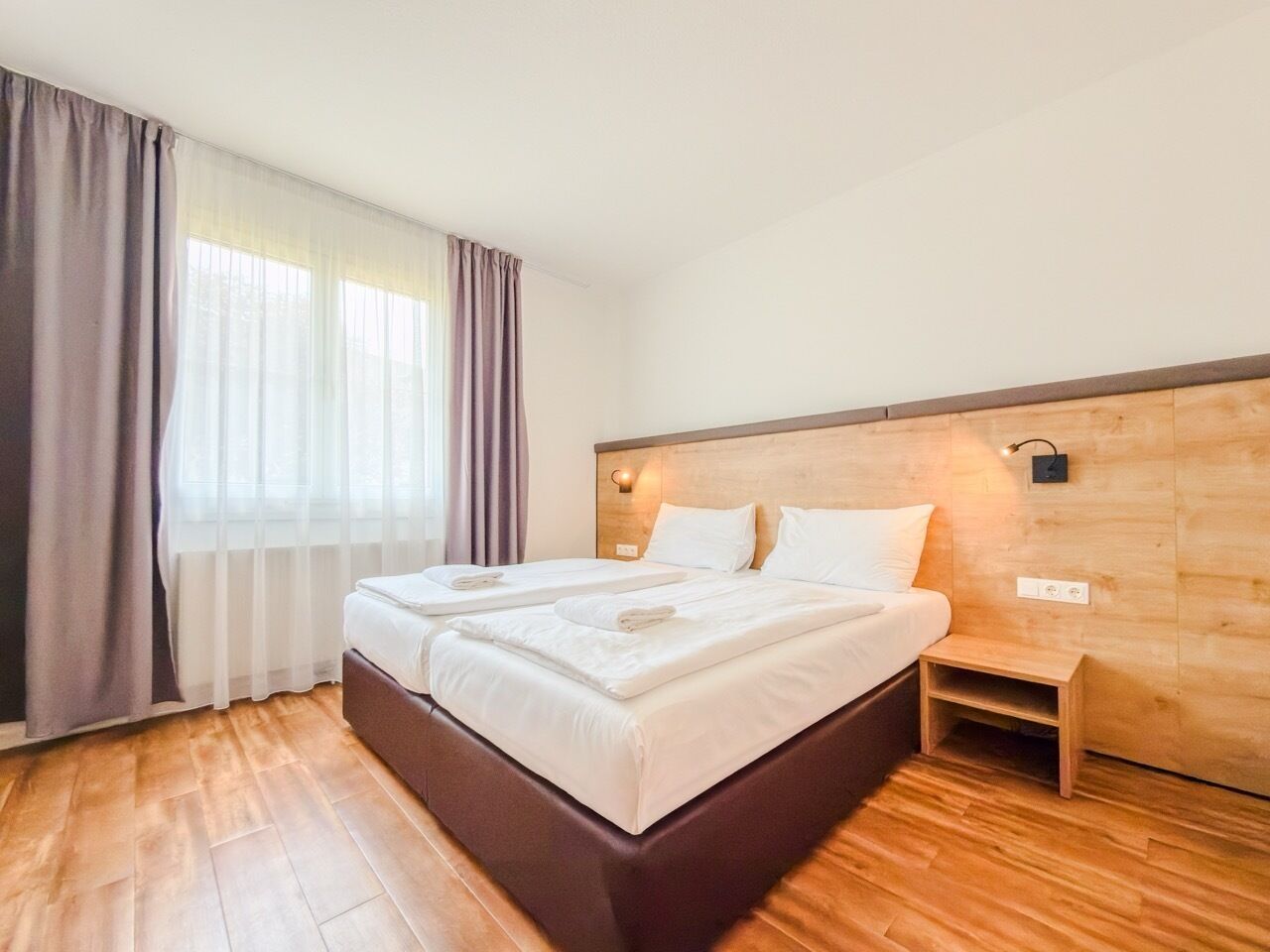 A double bed with a wooden headboard, a bedside table, and a window with curtains.