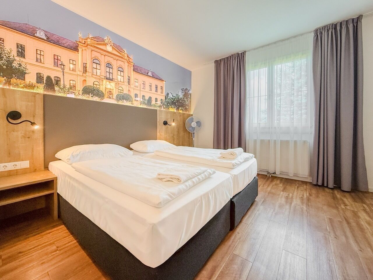 A hotel room with two beds, a mural of a building, a wooden desk, a fan, and a window with curtains.