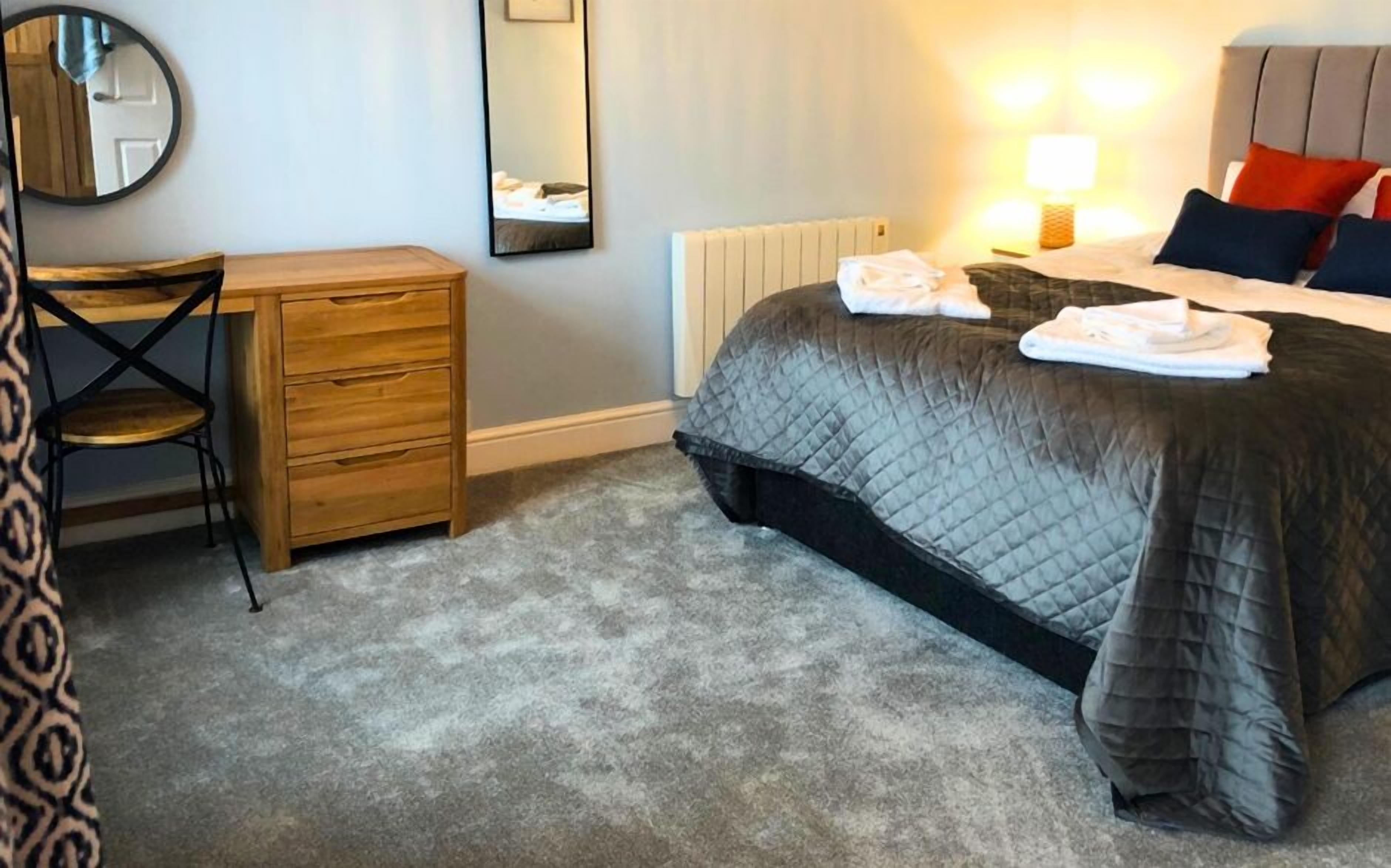 A hotel room with a bed, a bedside table, a chair, a dresser, a mirror, and a radiator.