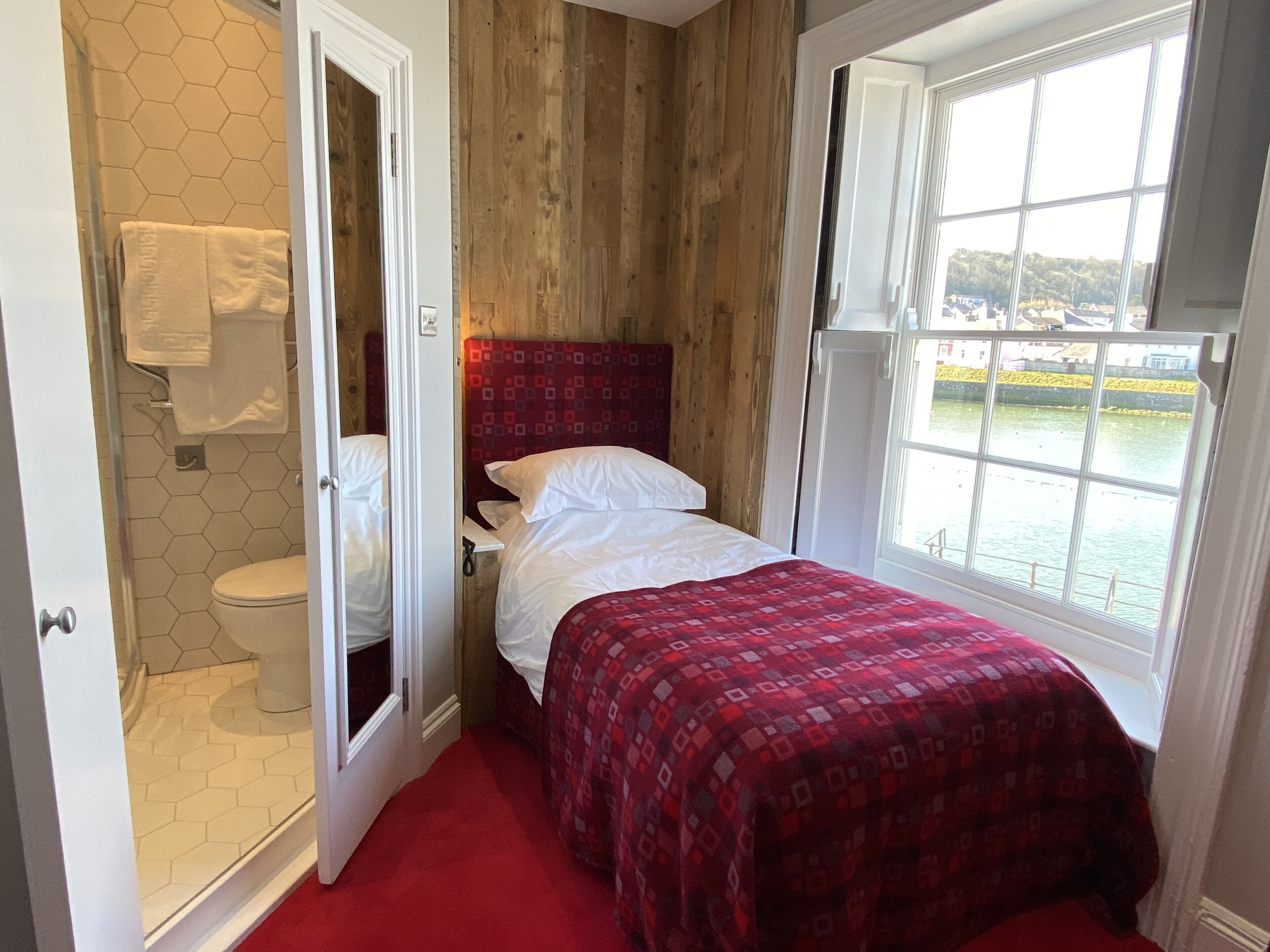 A bedroom with a wooden wall, a bed with a red patterned bedspread, a white headboard, and a window with a view of a body of water.