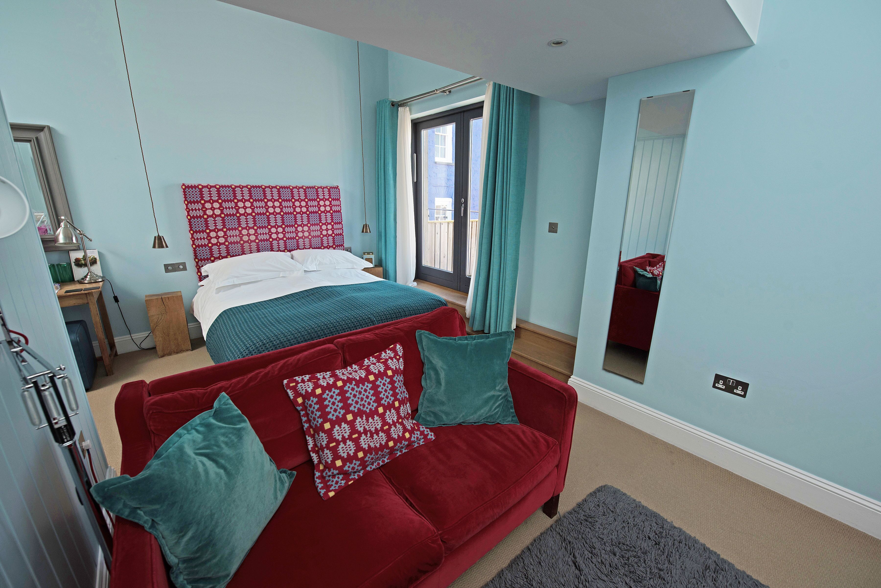A bedroom with a bed, a red sofa, a mirror, and a window with curtains.