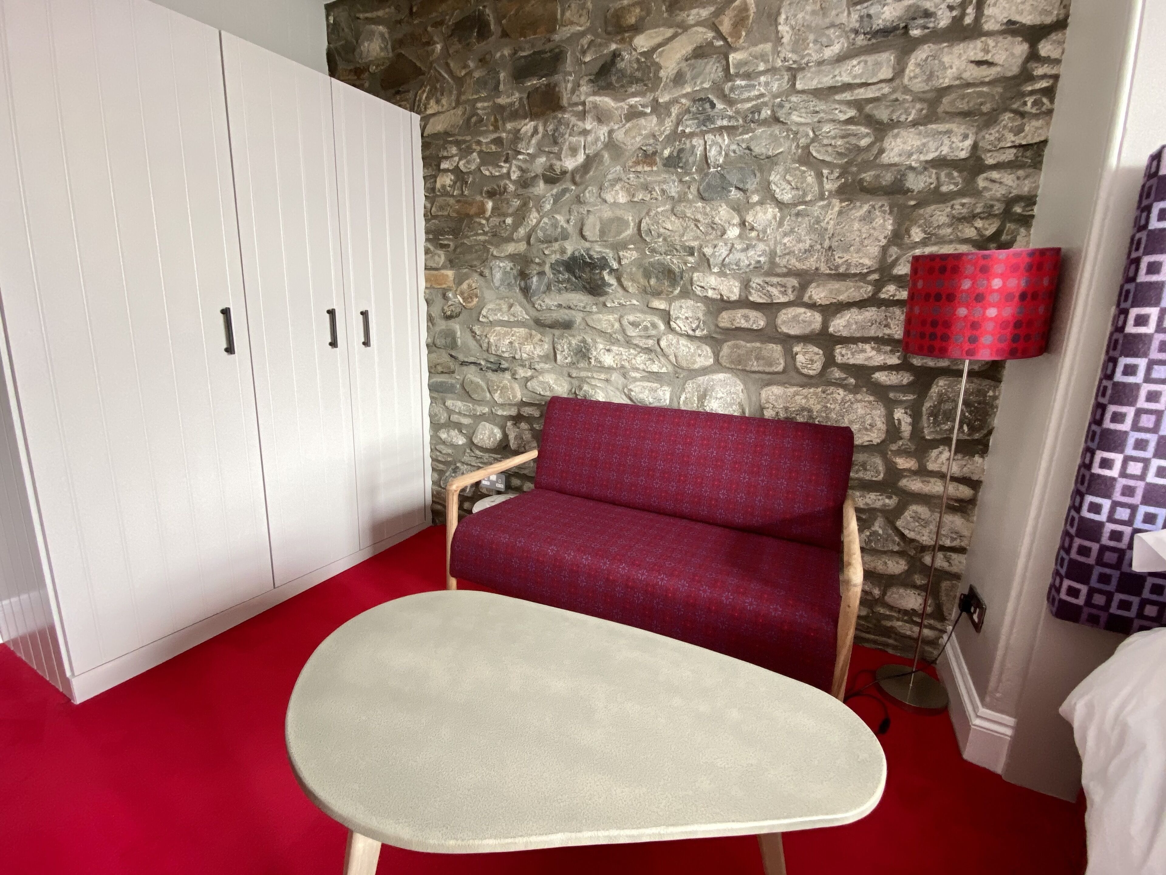 Una habitación con pared de piedra, un armario blanco, una silla roja y una mesita.