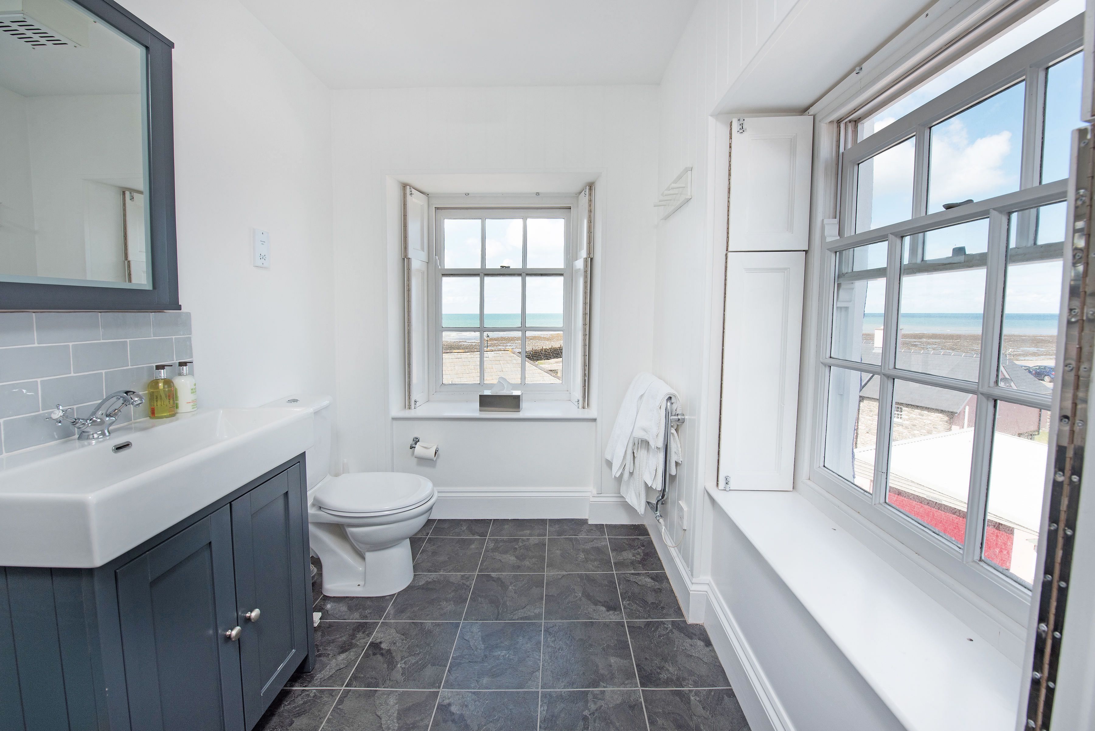 Un baño con lavamanos blanco, gabinetes gris oscuro, inodoro blanco y ventana con vista a la playa.