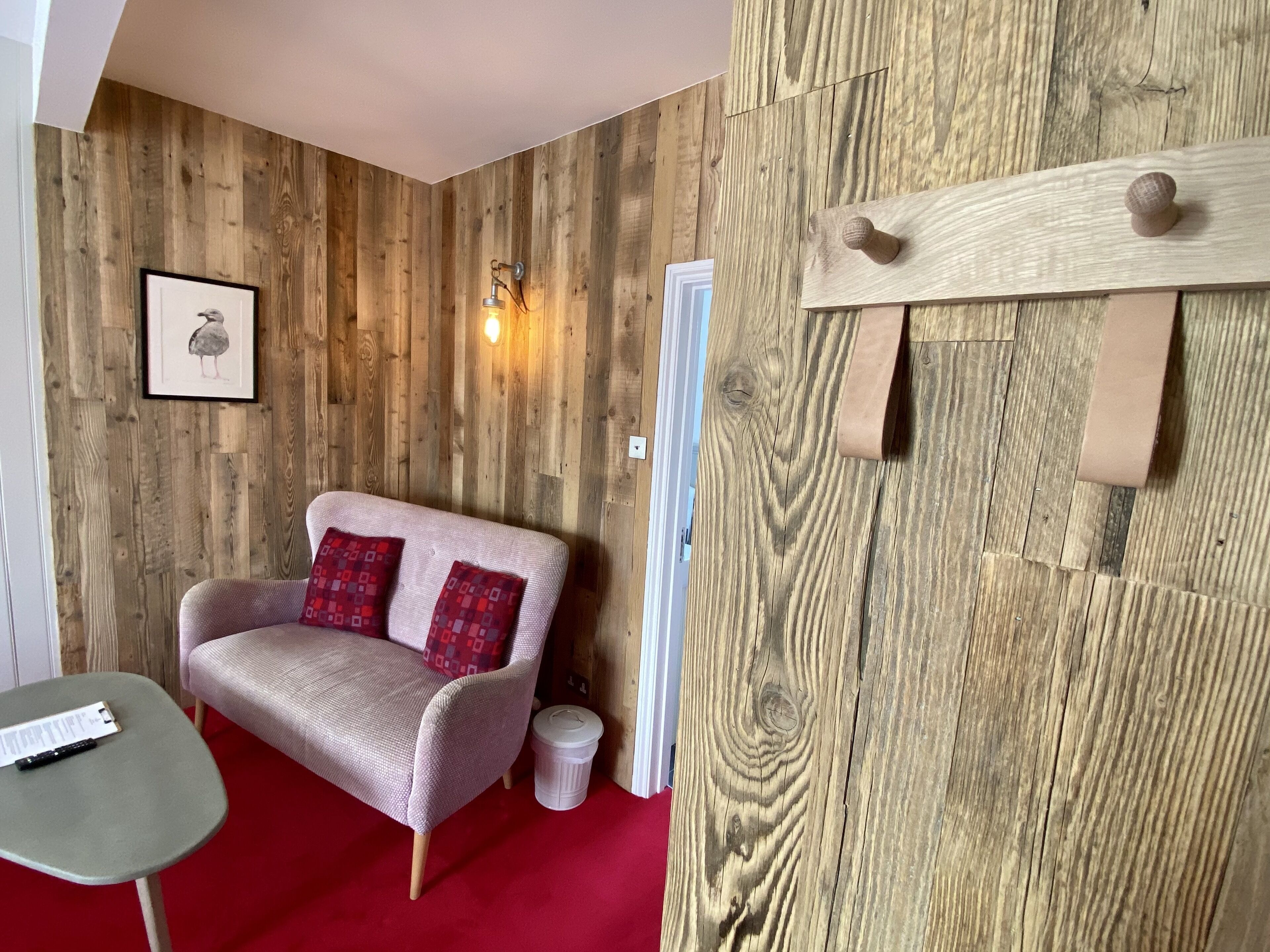 Una habitación con pared de madera, un sofá con cojines rojos, una mesita y un cubo de basura.