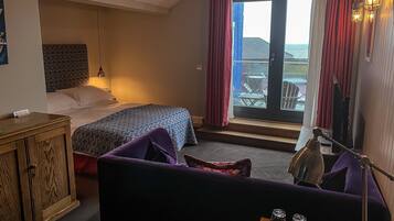 Ein Hotelzimmer mit Bett, Schreibtisch mit Lampe, violettem Sofa und Meerblick durch eine Schiebetür.