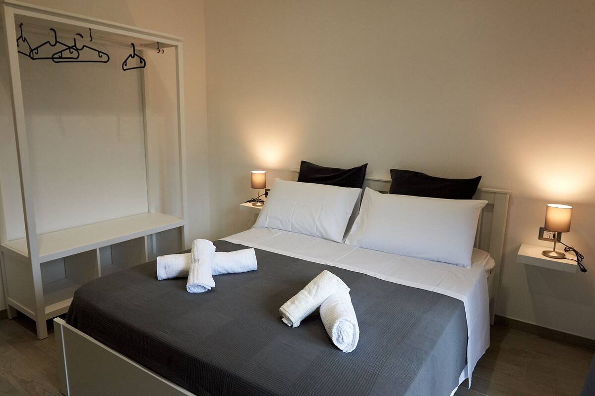 A neatly made bed with white linens and towels, bedside lamps, a wardrobe with hangers, and a bedside table.