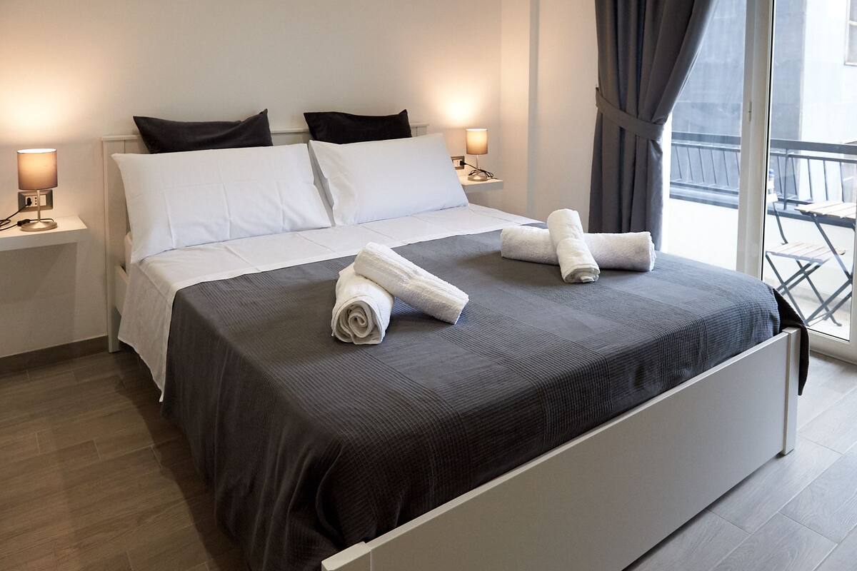 A neatly made bed with white and gray linens, two bedside lamps, and a view of the balcony through a window.