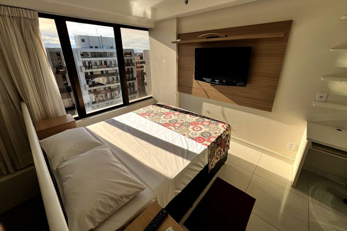 Une chambre d’hôtel avec un lit, un téléviseur fixé au mur, une fenêtre avec des rideaux et une vue sur les bâtiments environnants.