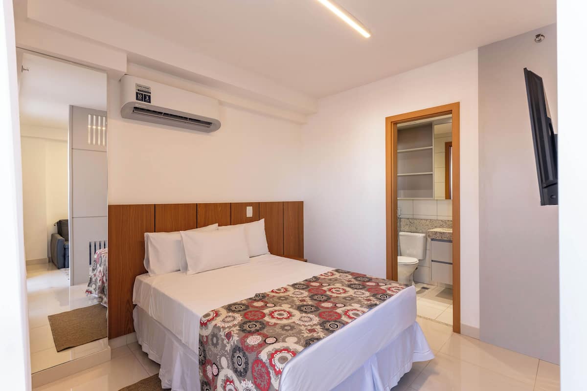 Une chambre d’hôtel avec un lit, une tête de lit en bois, une couvre-lit à motifs, un mur blanc, un climatiseur fixé au plafond et une salle de bain visible à travers une porte ouverte.