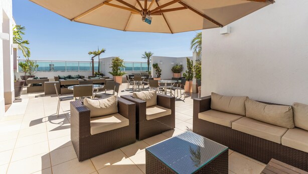 An outdoor patio with wicker furniture, a glass table, and a large umbrella. - Biarritz Hplus Long Stay (Brasília)