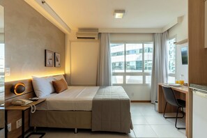 A hotel room with a bed, bedside table with a phone, a desk with a chair, a window with curtains, and an air conditioning unit. - Biarritz Hplus Long Stay (Brasília)