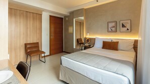 A hotel room with a bed, a desk with a chair, a wooden wardrobe, and a mirror. - Biarritz Hplus Long Stay (Brasília)