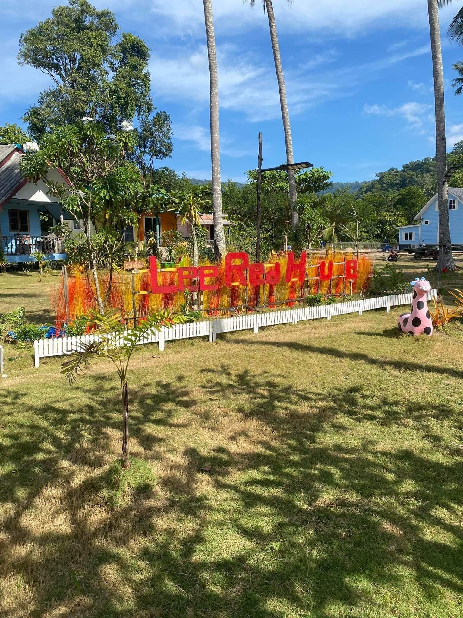 A sign with 'LEE REAL HOUSE' in red letters, surrounded by a white picket fence, palm trees, and houses in a sunny outdoor setting.