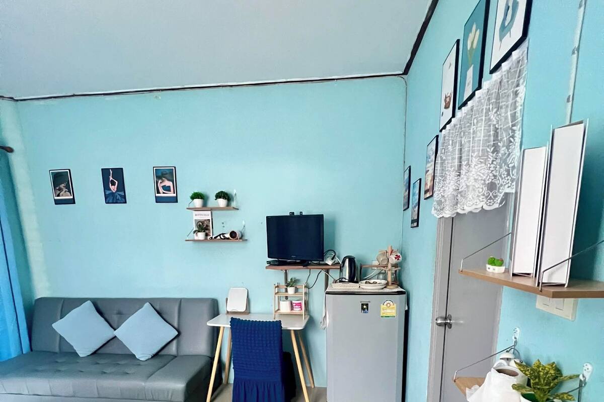 A compact living space with a sofa, a small table, a TV, a refrigerator, and shelves with plants and pictures.
