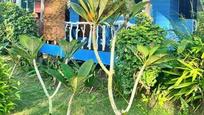 A house with a blue exterior, a porch with a white railing, and various tropical plants in the foreground. - Lee Red House (Ko Kood)