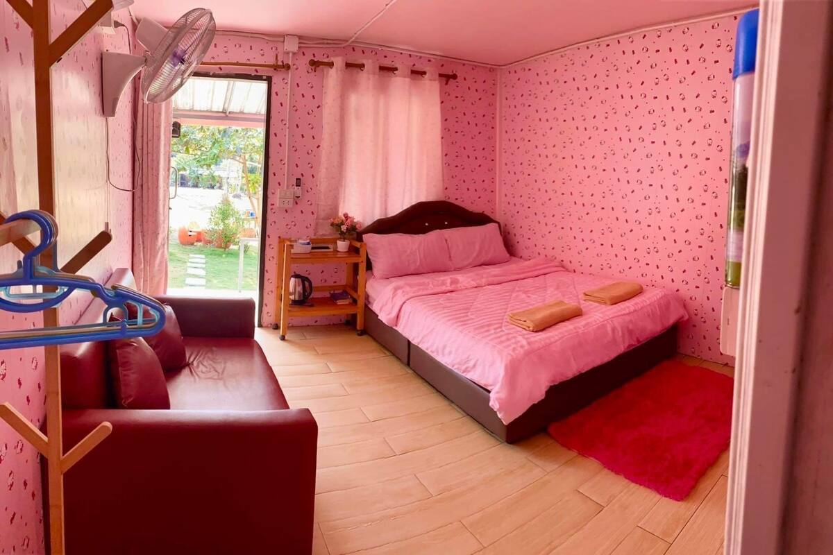 A bedroom with a bed, a wooden nightstand, a fan, a window with curtains, and a red rug.
