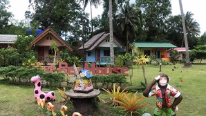 A garden with a windmill, a monkey statue, and a giraffe statue. There are also houses and a sign that reads "Real House". - Lee Red House (Ko Kood)