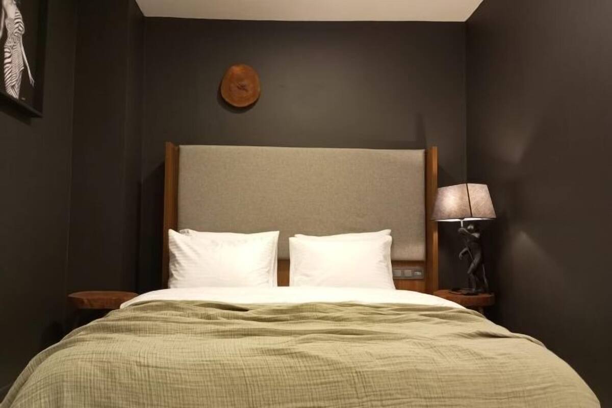 A hotel room with a bed, two pillows, a wooden headboard, a bedside lamp, and a wall-mounted artwork.