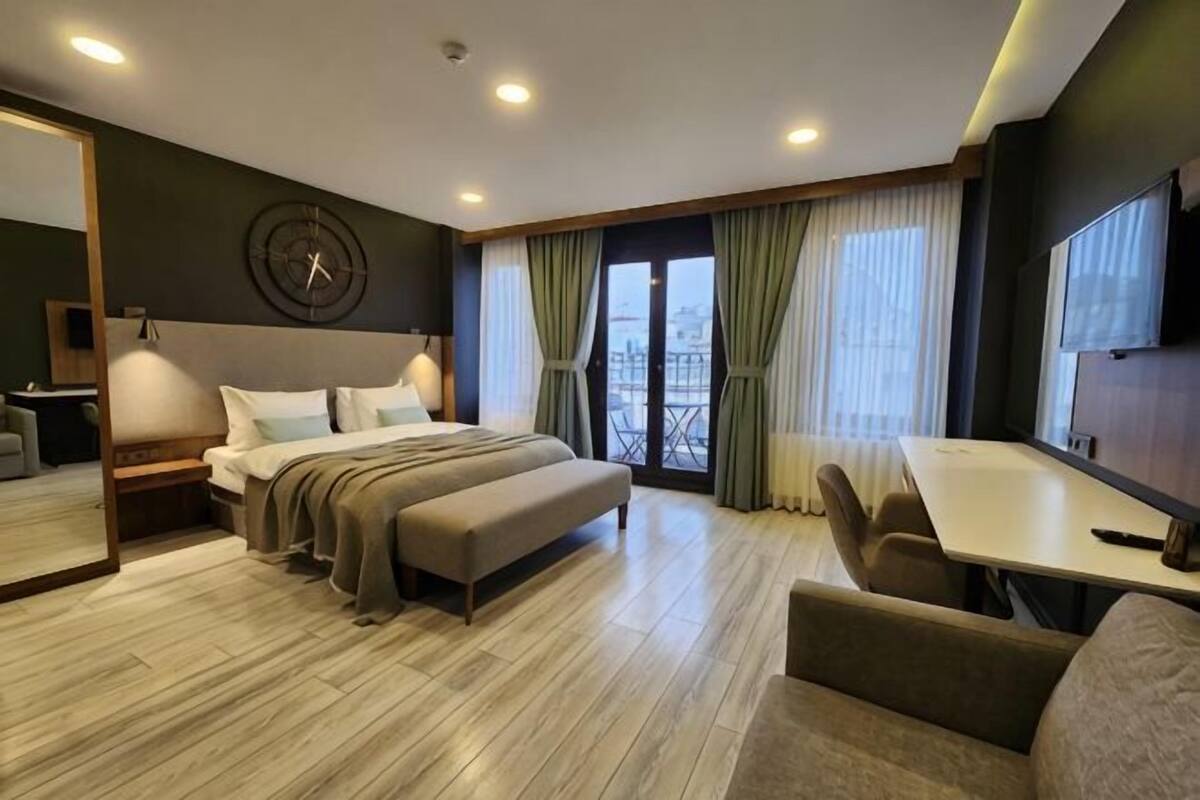 A hotel room with a bed, a desk, a chair, a bench, a TV, and a large window with curtains.