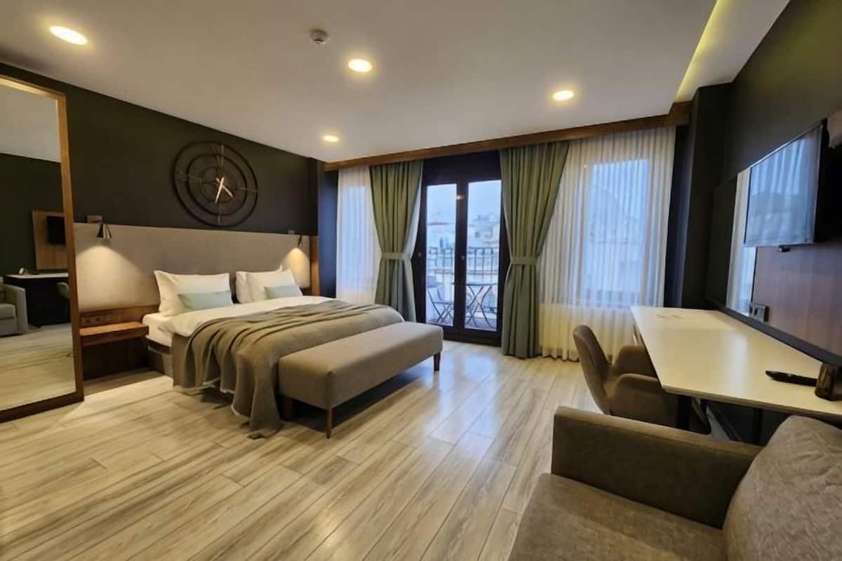 A hotel room with a bed, a desk, a chair, a bench, a TV, and a large window with curtains.