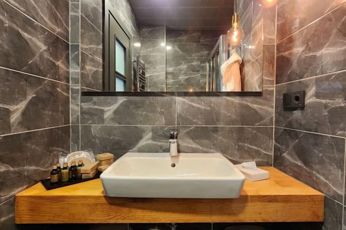 Un baño con pared de mármol, lavamanos blanco, tocador de madera y un espejo sobre el lavamanos.