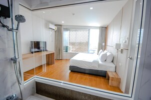 Habitación de hotel moderna con cabina de ducha de vidrio, cama con ropa blanca, suelo de madera y un amplio ventanal con vistas.