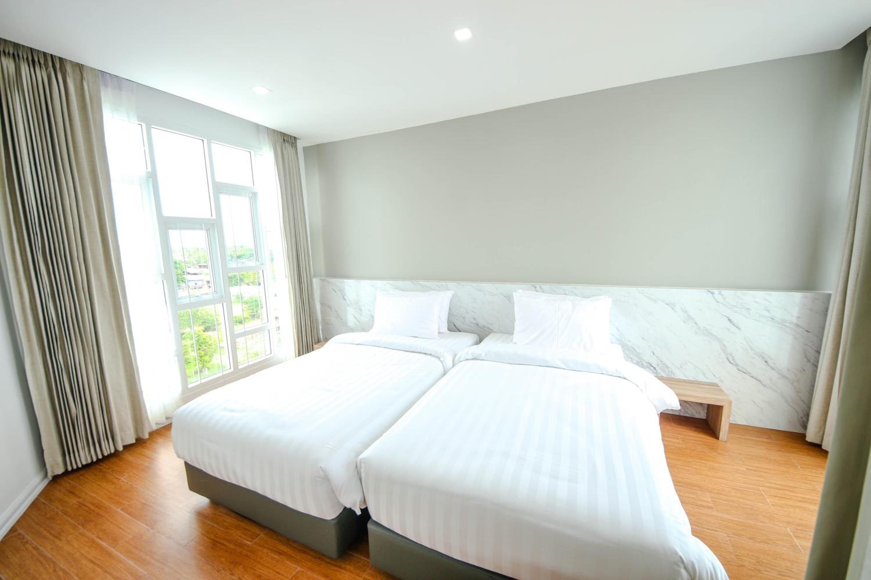 A hotel room with two beds, a marble headboard, wooden flooring, and large windows with curtains.