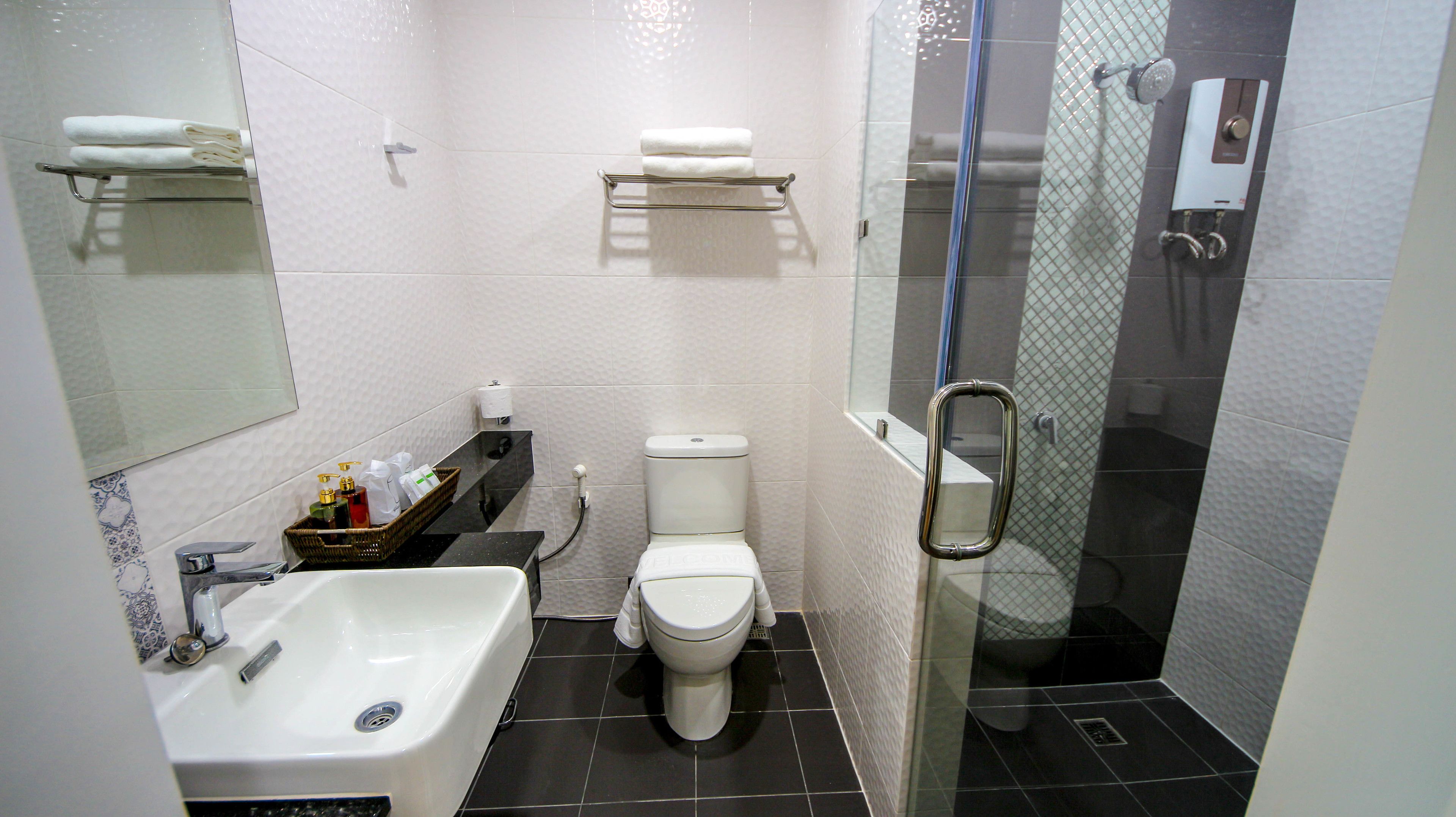 Un baño moderno con lavamanos, inodoro y ducha con mampara de vidrio. Hay toalleros sobre el inodoro y una pequeña cesta de artículos de aseo en la encimera.