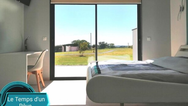 A bedroom with a bed, a desk, and a chair. There is a sliding glass door leading to a view of a grassy area. - Le Temps d'un Reve (Eauze)