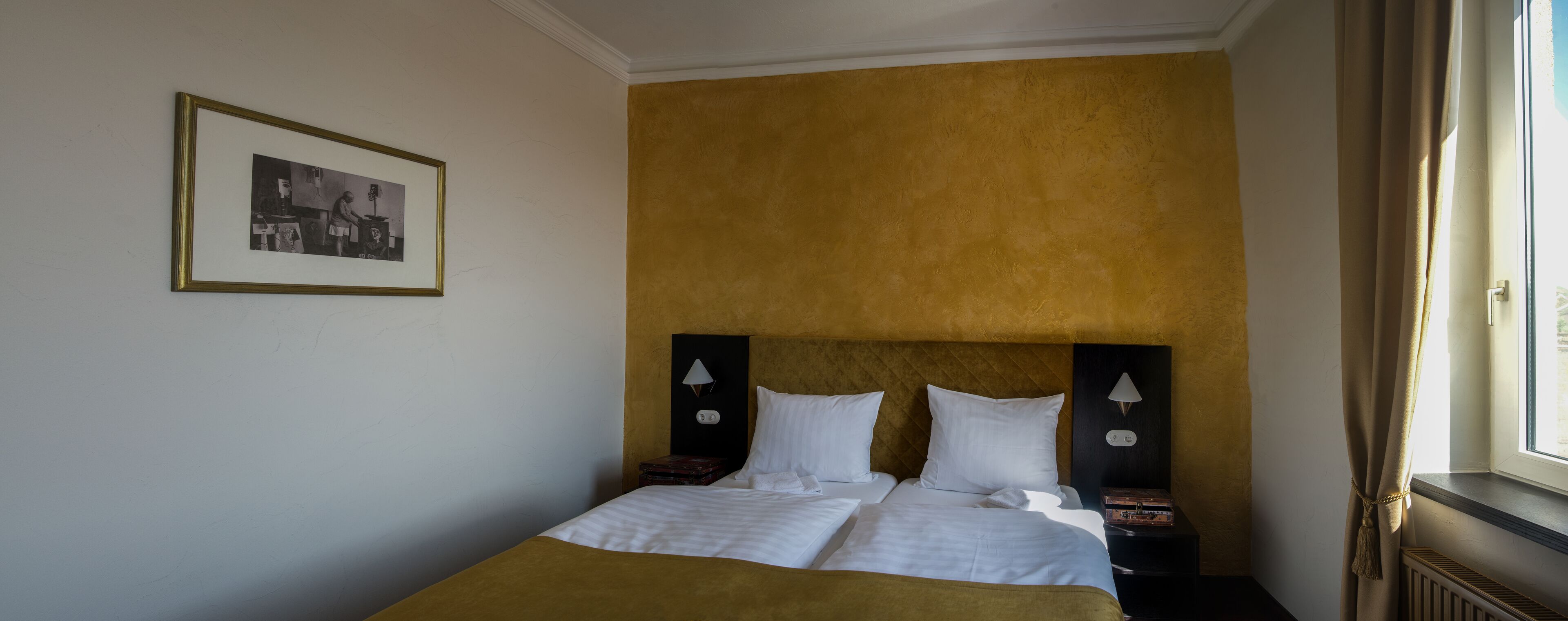 Une chambre d’hôtel avec un lit, deux tables de chevet, une commode, un tableau encadré au mur et une fenêtre avec des rideaux.