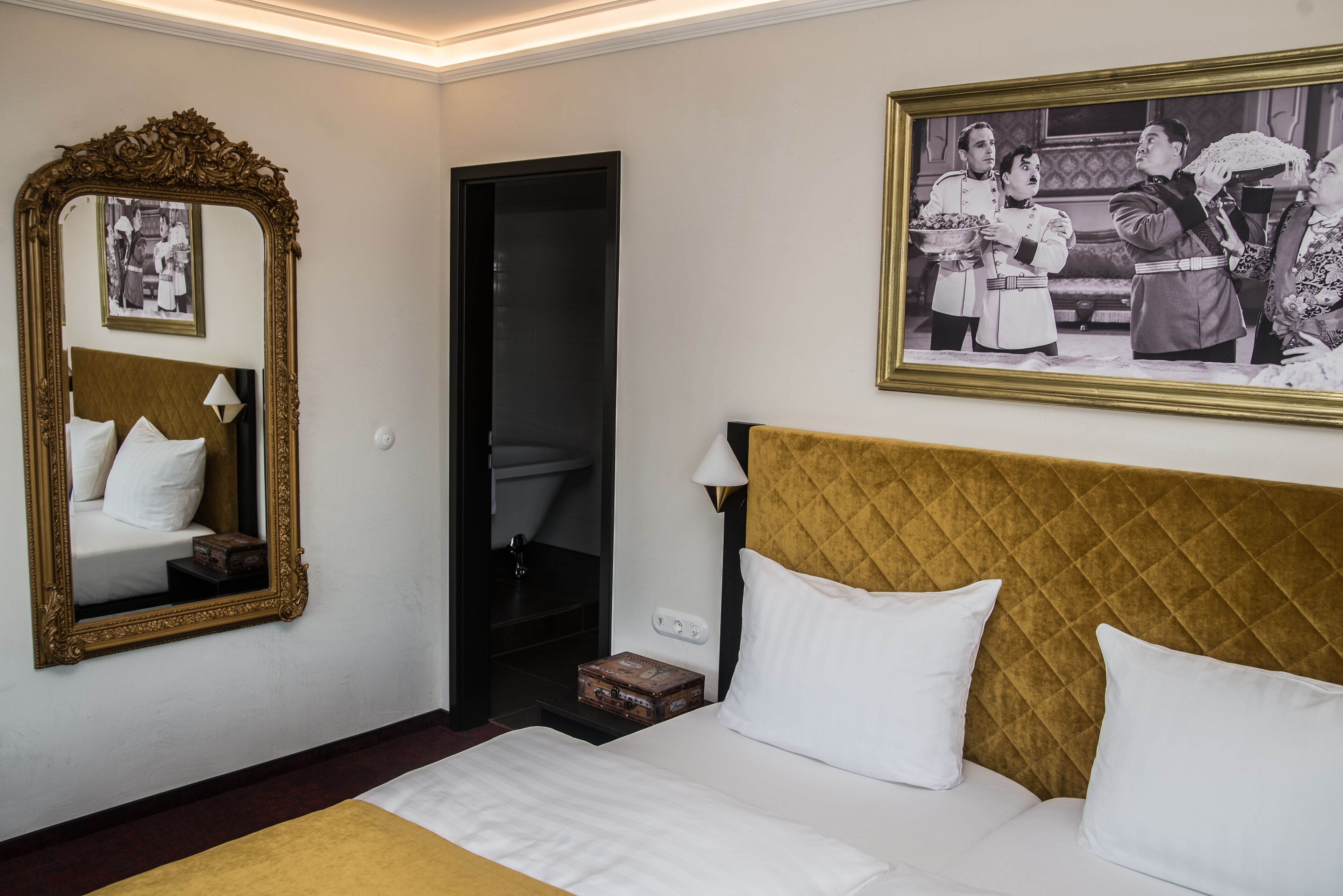 A hotel room with a large bed, a mirror, a framed picture on the wall, and a small table with a lamp.