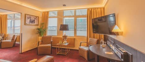 Habitación de hotel con alfombra roja, sofás de cuero, televisor, lámpara y ventana con cortinas.
