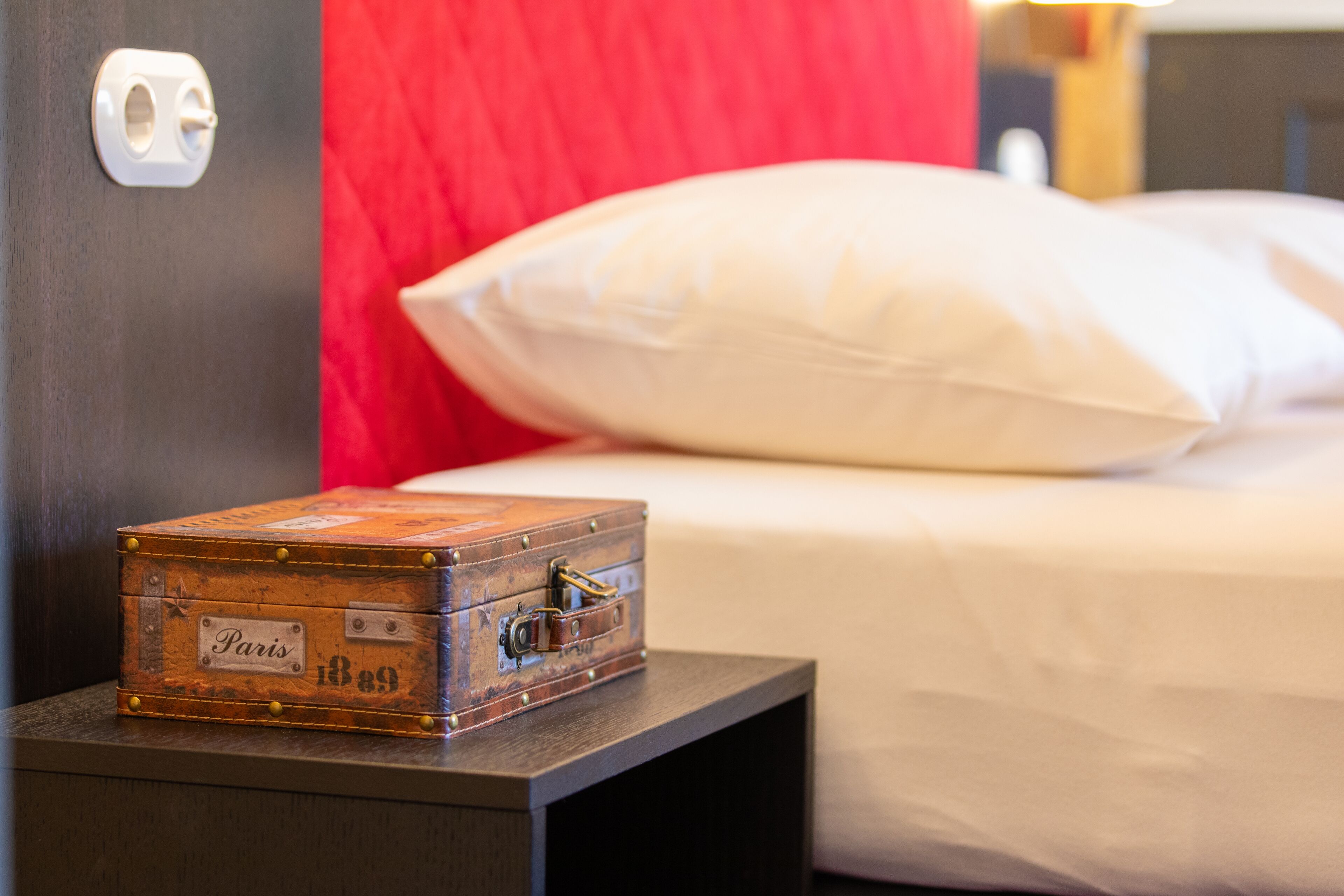 Een houten koffer met een label waarop 'Parijs 1889' staat, op een nachtkastje naast een bed met wit beddengoed en een rood hoofdeinde.